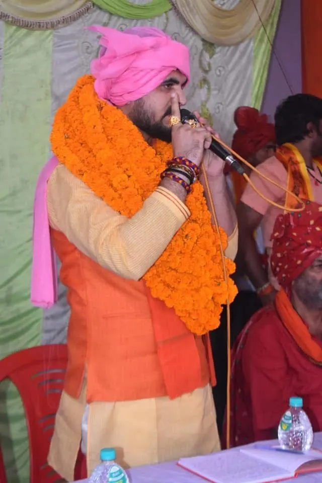 मंच पर माला पहने माइक्रोफ़ोन पकड़े वक्ता।